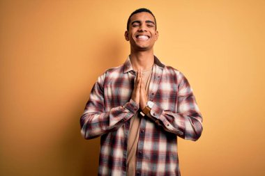 Genç, yakışıklı, Afro-Amerikan bir adam, günlük gömleğini giymiş sarı arka planda duruyor, ellerini birleştirip af diliyor, kendinden emin bir şekilde gülümsüyor..