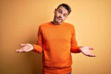 Sarı arka plan üzerinde duran turuncu günlük süveter giyen genç ve kafası karışmış, elleri ve kolları havada bir adam. Şüphe kavramı.