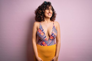 Genç, güzel bir Arap kadın, mayo ve pembe arka planda güneş gözlüğü takarak tatilde. Yüzünde gülümseme ve doğal bir ifadeyle diğer tarafa bakıyor. Kendinden emin gülen.