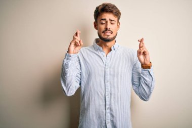 Sakallı, çizgili tişörtlü, beyaz arka planda duran yakışıklı genç adam umut ve gözler kapalı bir şekilde gülümseyerek işaret ediyor. Şans ve batıl inanç..
