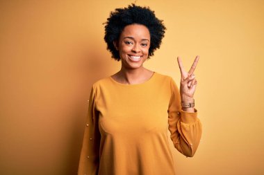 Kıvırcık saçlı, sıradan tişörtlü, kameraya gülümseyen ve zafer işareti yapan güzel Afro-Amerikan kadın. İki numara..