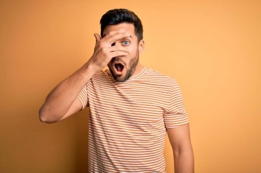 Sakallı, sarı arka planda sıradan çizgili tişört giyen genç yakışıklı bir adam. Şok olmuş bir şekilde yüzünü ve gözlerini gizliyor. Utanmış bir ifadeyle parmaklarının arasından bakıyor..