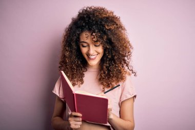 Kıvırcık saçlı ve pembe arka planda kitap okuyan, kendine güvenen gülümseyen ve gülümseyen güzel bir öğrenci kadın.