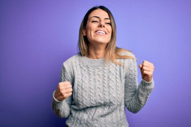 İzole edilmiş mor arka plan üzerinde sıradan bir süveter giyen genç ve güzel bir kadın mutlu ve heyecanlı bir şekilde kollarını kaldırıp, gülümseyerek ve başarı için çığlık atarak kazanan bir jest yapıyor. Kutlama konsepti.