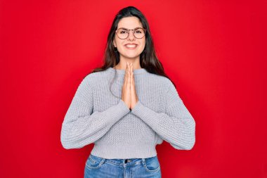 Genç, güzel esmer kadın, kırmızı arka plan üzerine gündelik süveter giyip ellerinin altında dua ederek af diliyor kendinden emin bir şekilde gülümseyerek..