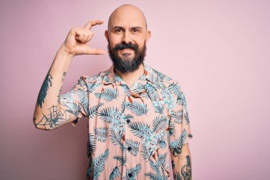 Sakallı ve dövmeli yakışıklı bir adam pembe arka planda sıradan bir çiçek gömleği giyiyor gülümsüyor ve el hareketleriyle el hareketleriyle küçük boy işareti yapıyor ve kameraya bakıyor. Ölçü kavramı.