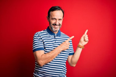 Orta yaşlı, beyaz, sıradan çizgili polo giyen, izole edilmiş kırmızı arka planda duran, gülümseyen ve kameraya bakan, iki eliyle ve parmaklarıyla yüzünü gösteren bir adam..