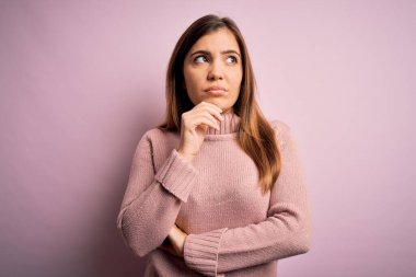 Güzel genç bir kadın pembe izole edilmiş arka plan üzerine balıkçı yaka kazak giyiyor ve elleri çenesinde soru sormayı düşünüyor, düşünceli bir ifade. Düşünceli bir yüzle gülümsüyor. Şüphe kavramı.