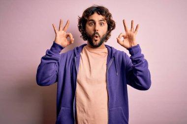 Sakallı, pembe arka planda sıradan bir kazak giyen yakışıklı genç bir adam. Şaşırmış ve şaşırmış bir şekilde parmaklarıyla onay sembolü çiziyor. Çılgın ifade
