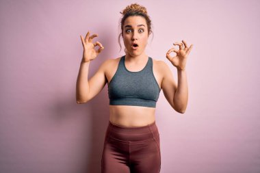 Pembe arka plan üzerine spor giyerek spor yapan genç ve güzel sarışın kadın. Şaşırmış ve şaşırmış bir şekilde parmaklarıyla onay sembolü çiziyor. Çılgın ifade