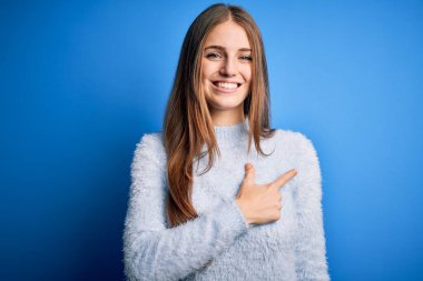 Genç, güzel, kızıl saçlı bir kadın, izole edilmiş mavi arka plan üzerine günlük süveter giyiyor. Neşeli, yüzünde mutlu ve doğal bir ifadeyle el ve parmağı gösteren bir gülümseme var.