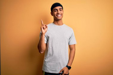 Yakışıklı Afro-Amerikan sporcu sarı arka planda spor giysisi giyiyor yüzünde kocaman bir gülümsemeyle kameraya el ile işaret ediyor..