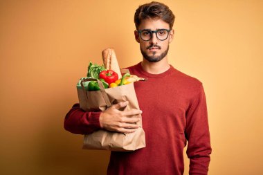 Gözlük takan genç bir adam elinde yiyecek dolu bir torba ile izole edilmiş sarı bir arka plan ve akıllı bir yüz ifadesiyle ciddi ciddi düşünüyor.