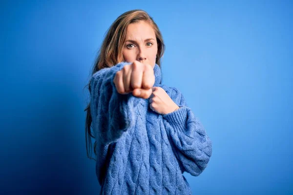 若いです美しいですブロンド女性身に着けていますカジュアルTurtleeckセーター上の青の背景パンチ拳へ戦います 積極的かつ怒っている攻撃 脅威と暴力 — ストック写真