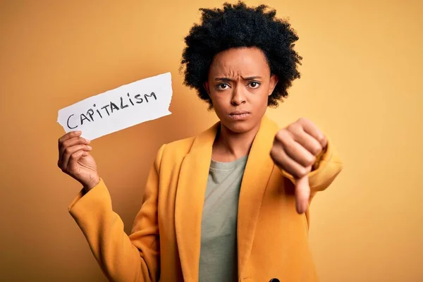 アフリカ系アメリカ人のアフロビジネス女性で 資本主義のメッセージと怒りの顔を持つ巻き毛保持紙 親指で嫌いを示す負の記号 拒否の概念 — ストック写真