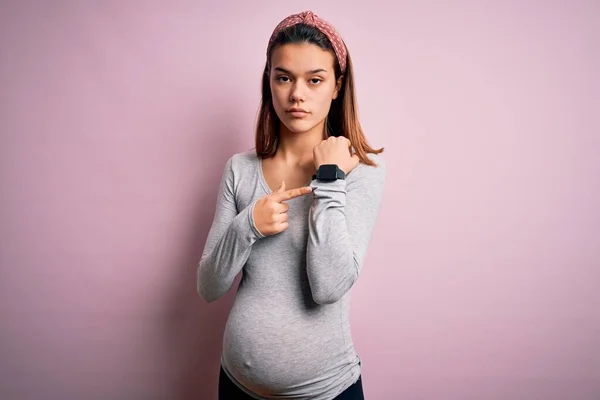 若いです美しいですティーン女の子妊娠期待赤ちゃん上の隔離されたピンクの背景で急いで時間を見て リラックスした表情でカメラを見て — ストック写真