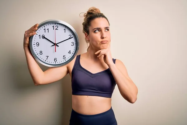 Young beautiful blonde sportswoman wearing sportswear on time to do sport holding clock serious face thinking about question, very confused idea
