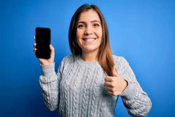 青い隔離された背景の上に空白のスマートフォンの画面を示す若い女性幸せな大きな笑顔で大丈夫サインをします 指で親指アップ 優れたサイン — ストック写真