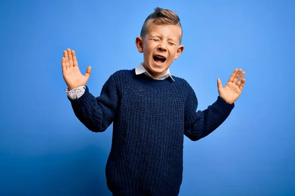 青い背景に冬のセーターを着た青い目の若い白人の子供は 腕を上げて興奮して叫んで目を閉じて成功のために狂気と狂気を祝います 勝者のコンセプト — ストック写真
