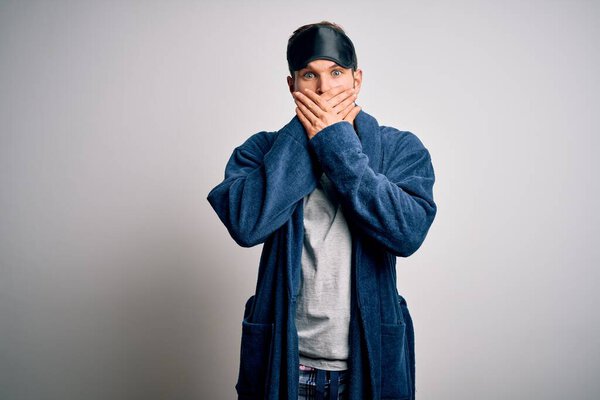 Young blond man with beard and blue eyes wearing sleep mask and pajama shocked covering mouth with hands for mistake. Secret concept.