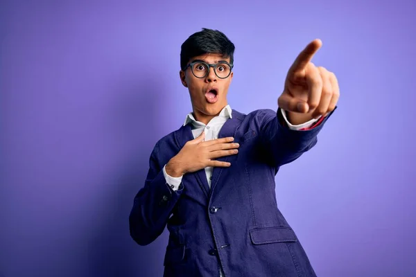 Young handsome business man wearing jacket and glasses over isolated purple background Pointing with finger surprised ahead, open mouth amazed expression, something on the front