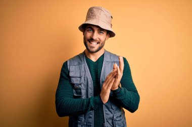 Tatilde sakallı yakışıklı turist sarı arka planda kaşif şapkası takıyor alkışlıyor ve mutlu ve neşeli, gururlu elleri alkışlıyor.