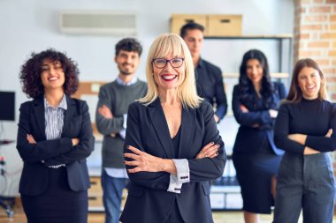 Bir grup iş adamı mutlu ve kendinden emin gülümsüyor. Yüzünde tebessümle birlikte kameraya bakarak poz vermek, ofiste kollarını kavuşturmuş orta yaşlı güzel bir kadın.
