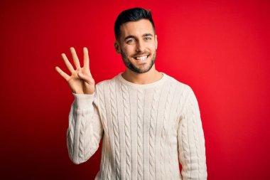 İzole edilmiş kırmızı arka plan üzerinde duran, rahat beyaz süveter giyen genç ve dört numaralı parmaklarıyla işaret eden, kendinden emin ve mutlu gülümseyen bir adam..