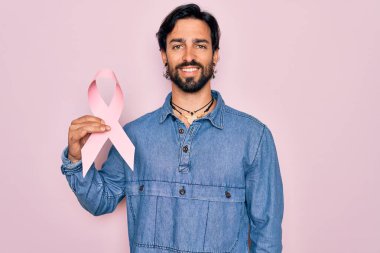 Göğüs kanseri tutan genç yakışıklı İspanyol adam mutlu yüzlü pembe kurdeleli farkındalık dişlerini gösteren kendine güvenen bir gülümsemeyle ayakta duruyor.