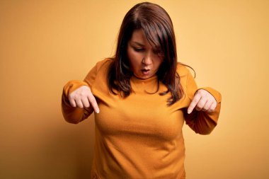 Güzel esmer ve cüsseli bir kadın izole edilmiş sarı arka plan üzerine gündelik süveter giyip parmaklarını göstererek reklamını yapıyor, şaşırmış yüzünü ve ağzını açıyor.