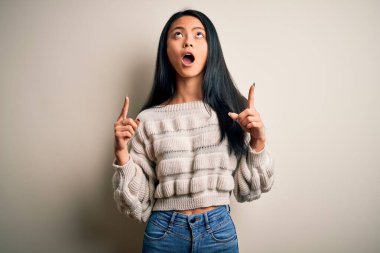 İzole edilmiş beyaz arka plan üzerine gündelik süveter giyen genç güzel Çinli kadın şaşırmış ve şaşırmış bir şekilde yukarı bakıp parmaklarını göstermiş ve kollarını kaldırmıştı..