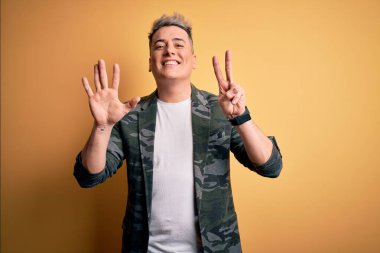 Sarı arka planda duran iş ceketi giyen yakışıklı ve modern bir adam yedinci parmaklarını gösterirken kendinden emin ve mutlu bir şekilde gülümsüyor..