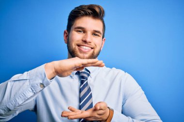 Sakallı, mavi gözlü, zarif gömlekli ve kravatlı genç sarışın işadamı. Büyük ve büyük beden işareti gösteren hareketlerle ayakta duruyor. Ölçü sembolü. Kameraya bakıp gülümsüyor. Ölçüm konsepti.