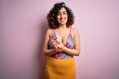 Tatilde olan genç, güzel bir Arap kadın. Mayo ve pembe arka planda güneş gözlüğü takıyor. Elleri çapraz, rahat ve neşeli bir şekilde gülümsüyor. Başarı ve iyimser
