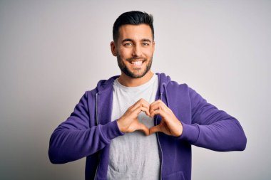 Mor kazak giyen yakışıklı genç adam izole edilmiş beyaz arka planda duruyor. Aşkla gülümsüyor. Elleriyle kalp sembolü ve şekli gösteriyor. Romantik konsept.
