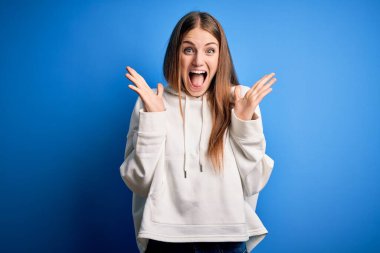 Genç, güzel, kızıl saçlı sporcu kadın mavi arka plan üzerine eşofman giyip çılgınlığı kutluyor ve kollarını kaldırıp heyecanla haykırarak başarıyı kutluyor. Kazanan konsept