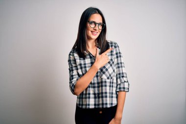 Mavi gözlü genç esmer kadın, günlük gömlek ve gözlük takıyor, beyaz arka planda neşeli, yüzünde gülümseme, eli ve parmağı yanında mutlu ve doğal bir ifade var.