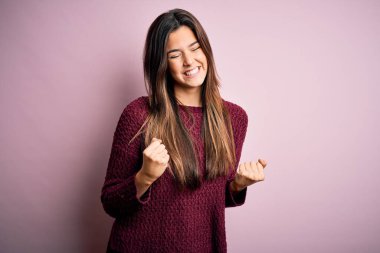 İzole edilmiş pembe arka plan üzerine sıradan bir süveter giyen genç ve güzel bir kız çok mutlu ve heyecanlı bir şekilde kollarını kaldırıp, gülümseyerek ve başarı için çığlık atarak kazanan bir jest yapıyor. Kutlama konsepti.