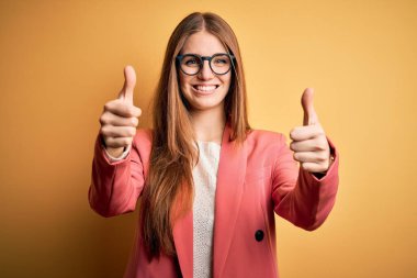 Ceket ve gözlük takan genç, güzel, kızıl saçlı bir kadın. Sarı arka planda el hareketlerini onaylıyor. Başparmağıyla gülümsüyor ve başarı için seviniyor. Kazanan jest.