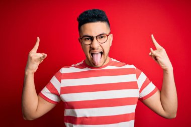 Sıradan çizgili tişört ve gözlük takan genç yakışıklı adam izole edilmiş kırmızı arka planda çılgın bir ifadeyle elleri havada rock sembolü yaparak bağırıyor. Müzik yıldızı. Ağır müzik konsepti.