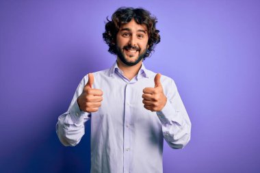 Mor arka plan başarısı tabelasının üzerinde duran sakallı, sakallı genç ve yakışıklı bir iş adamı. Elleriyle olumlu bir jest yapıyor. Gülümsüyor ve mutlu. Neşeli ifade ve kazanan jest.