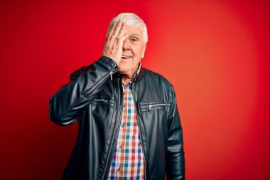 Kıdemli yakışıklı, beyaz gömlekli ve ceketli bir adam izole edilmiş kırmızı arka planda bir gözünü elinle kapatıyor, yüzünde kendine güvenen bir gülümseme ve sürpriz bir duygu var..