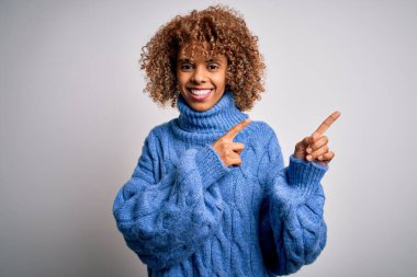 Beyaz arka planda boğazlı kazak giyen, gülümseyen ve kameraya bakıp iki eliyle ve parmaklarıyla yüzünü gösteren güzel Afro-Amerikan kadını..