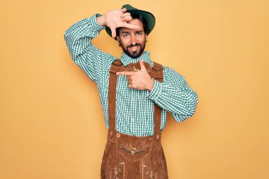 Almanya festivali için geleneksel Alman Ekim Festivali kıyafeti giyen yakışıklı genç adam gülümseyerek elleri ve parmakları gülen yüzle çerçeve yapıyor. Yaratıcılık ve fotoğrafçılık kavramı.