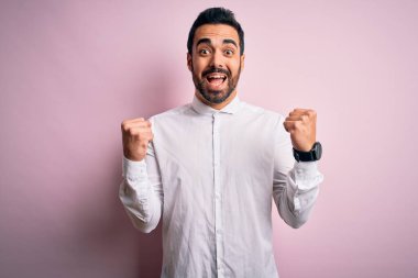 Young handsome man with beard wearing casual shirt standing over pink background celebrating surprised and amazed for success with arms raised and open eyes. Winner concept.