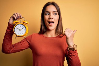 Sarı arka plan üzerinde çalar saat tutan genç, güzel esmer bir kadın. Gülümseyen bir gülümsemeyle baş parmağıyla işaret ediyor.