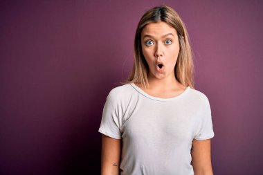 Genç, güzel, sarışın bir kadın mor bir arka plan üzerine günlük beyaz tişört giyiyor. Şaşırtıcı bir ifade, korku ve heyecanlı bir yüz ifadesiyle dehşete kapılmış ve şok olmuş..