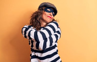 Orta yaşlı, esmer, maske takan, kep takan, sarı arka planda dikilen, mutlu, pozitif ve kendinden emin gülümseyen bir hırsız kadın. Sevgi ve özgüven.