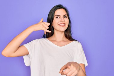 Genç, güzel, esmer bir kadın, mor arka plan üzerine günlük beyaz tişört giyip gülümsüyor, telefonda konuşuyor ve seni işaret ediyor. Ara beni..