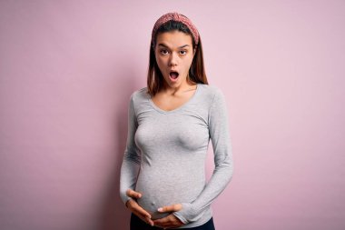Genç, güzel, hamile bir kız pembe arka planda bebek bekliyor. Şaşırmış ve şaşırmış bir ifade, korku ve heyecanlı bir yüz..